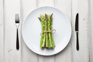 Dinner plate with bunch of fresh asparagus, fork and knife on white wooden background, top view