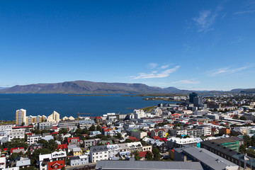 Aerial view on capital of Iceland - city of Reykjavik - ocean bay, hills, mountains, green medows, roadways, vehicles, streets, houses, traffic, roofing - on sunny day.
