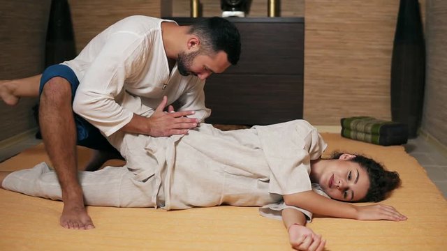 Male thai massagist is treating young woman's hip and sciatic nerve holding her leg and stretching them. Thai massage session for young attractive woman