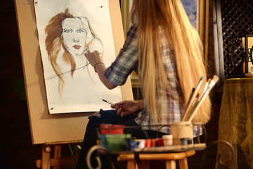 Artist painting on easel in studio. Girl paints portrait of woman with brush. Female painter seen from behind. Indoor home interior for handmade crafts. Student works as an artist.