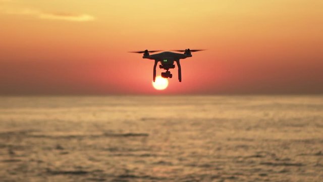 Silhouette of a flying drone over calm sea with captivating golden sunset and flying birds background slow motion
