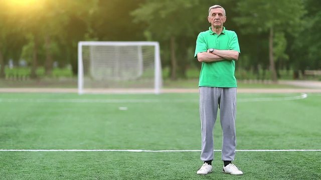 Football trainer stands on football field. Senior man in green t-shirt stands on soccer field and looks at camera, football gate and morning sun with flicker at background. Physical education teacher