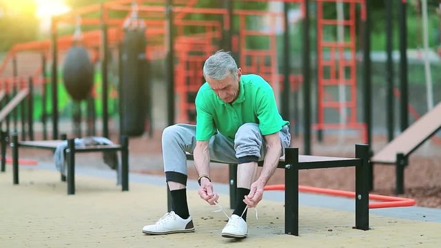 Senior man laces up his jogging shoes near sports ground. Grey-haired elderly athlete sits on the bench near outdoor gym and laces up his sneakers, beautiful morning sun with flicker at background