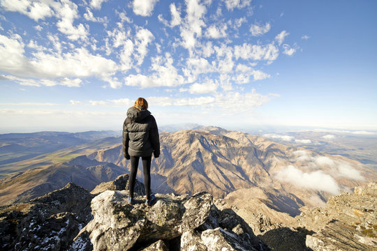 Red haired female hiker standing on the edge of a mountain overlooking a mountain top with clouds.