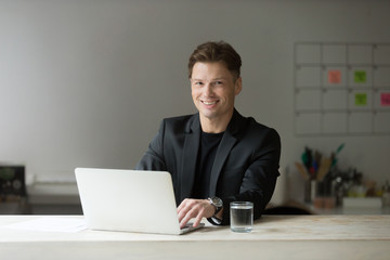 Portrait of handsome smiling businessman in suit in modern office in front of laptop, looking at camera. Positive business person during important meeting, being friendly to clients and coworkers.