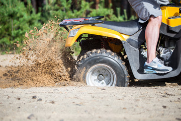 An ATV quadbike get stuck in a sandy road near forest and having wheel-spin making a spray of sand