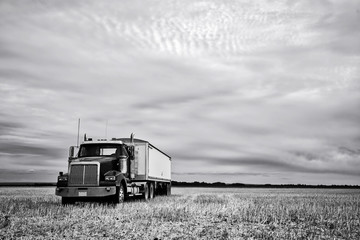 A semi truck with an attached grain trailer parked on a harvested field in a black and white countryside landscape