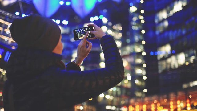 Woman Using SmartPhone at Night in Big City. 4K. Attractive young Woman taking photos on cellphone outdoors. Millennial Girl with phone, blurred Street skyscrapers and traffic lights.