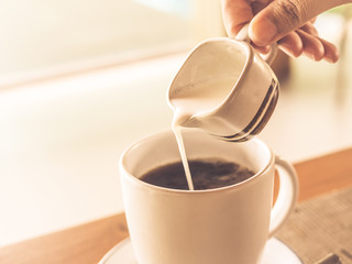 Closeup woman hand  pouring hot milk into black coffee cup during breakfast time, with selective focus.