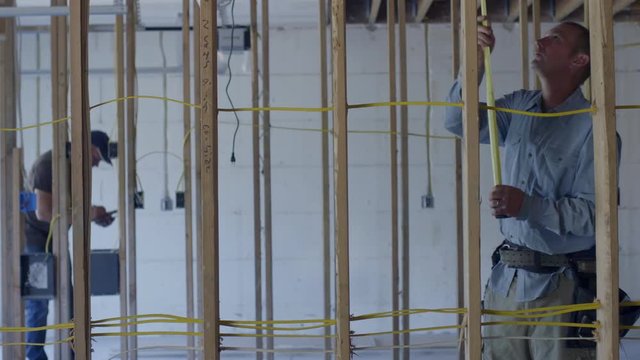 Contractor measures wooden beams while his colleague checks various items in background of partially finished basement garage. Medium long shot with slider move.