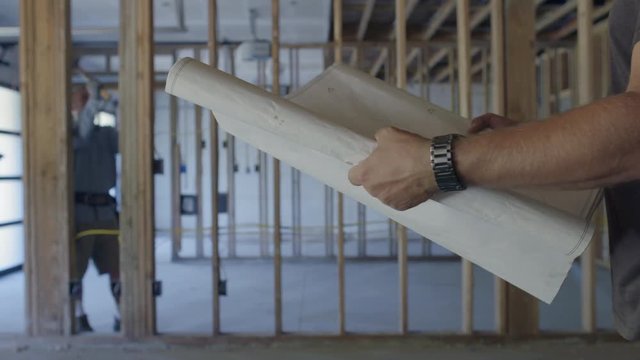 Workman hammers nails in partially finished basement garage while colleague examines plans in close up foreground. Side view with rack focus and slider move.