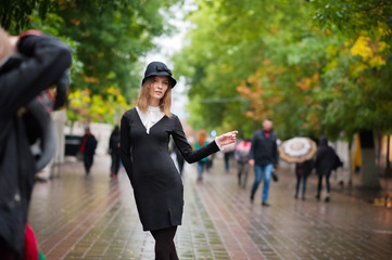 A beautiful thin girl in an amazing black dress in retro style on the background of a pedestrian street. A girl in a black hat after the rain. Emotion of the girl. Emotional face.