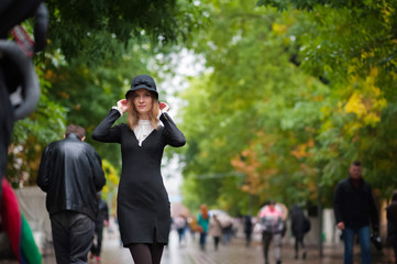 A beautiful thin girl in an amazing black dress in retro style on the background of a pedestrian street. A girl in a black hat after the rain. Emotion of the girl. Emotional face.