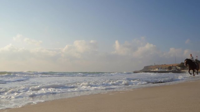 Beautiful young women riding dark horses at sea beach. Enjoying beautiful landscape.Galloping run in sunset or sunrise light and splashing water drops around.Riders running at a gallop.Slow motion.