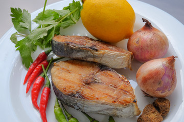 A grilled mackerel fish on white plate  with onion, chilli, lemon , belacan and soup leaf. A grilled mackerel fish  is a one of the Malaysian people favorite dish.