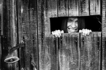 Portrait of a smiling woman peering through the crack in the barn door. Black-and-white photo.