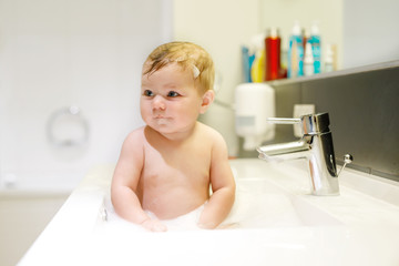 Cute adorable baby taking bath in washing sink and playing with water and foam