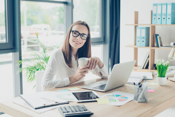 True happiness. Successful charming relaxed brown haired economist is enjoying beverage at her work station, in smart outfit, black trendy eyewear, serene, carefree, resting mode