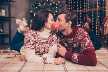 Closeup low angle of adorable dreamy sweet cute friends kissing behind tasty yummy treat with bow on stick, lying down on floor on plaid, carpet, winter feast, coziness, warm, in socks, pine tree