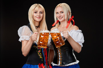 Two beautiful blonde women are holding glasses of beer in hands and stand on black background in studio.