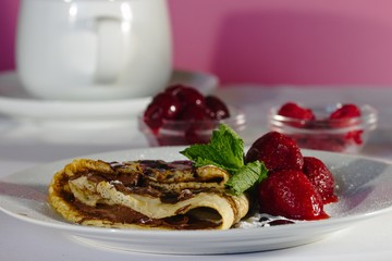 Serving of homemade crepes with strawberries, chocolate topping and powdered sugar. Small jars of red berries and teacup in background.