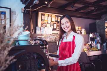 Obraz na płótnie Canvas Beautiful asian barista wearing red color apron working with coffee maker machine and smiling at camera