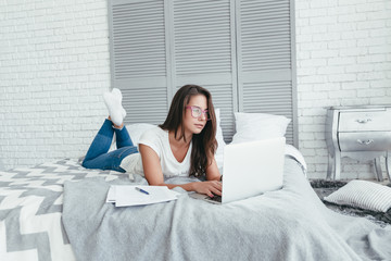 Beautiful young woman in glasses working from home on a laptop lying on the bed in the room