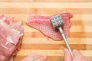 Chef's hands with a hammer beating a meat on the wooden board in the kitchen. Preparation for...