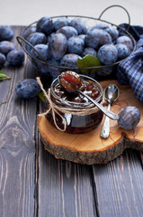 Homemade plum jam in a glass jar and fresh blue plums in a bowl on a dark rustic wooden background top view.