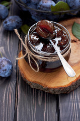 Homemade plum jam in a glass jar and fresh blue plums in a bowl on a dark rustic wooden background top view.