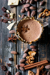 Vintage mug of hot chocolate, decor with nuts, caramel, spices. Ingredients above. Chopped dark and white chocolate, cocoa beans, anise over old wooden table. Top view with space. Dark rustic style