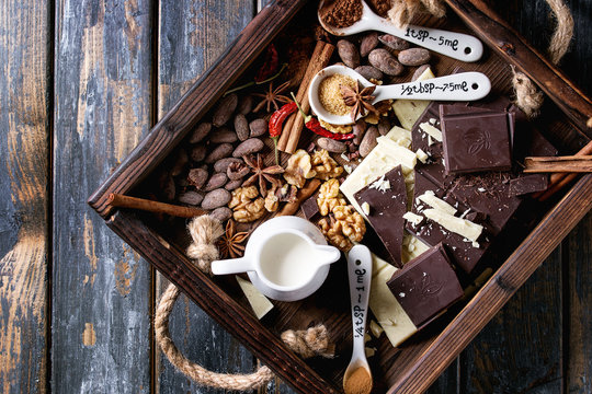 Ingredients for cooking hot chocolate. White and dark chopping chocolate, cocoa powder, cocoa beans, cream, cinnamon, sugar in spoons in wood tray over old wooden background. Top view with space