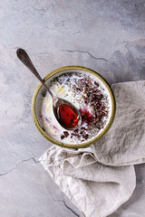 Bowl of milk cereal porridge with additives jam, chocolate, chia seeds and berries, served with spoon over gray kitchen table. Top view with space