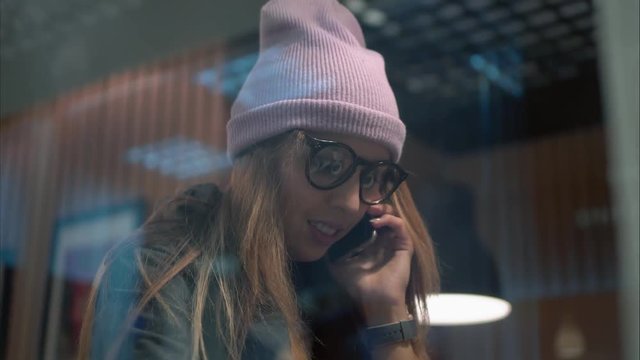 A stylish hipster female in glasses talking on phone drinking coffee in a cafe