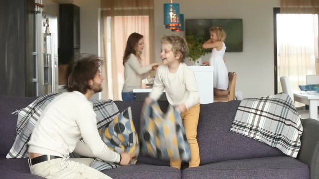 Happy family couple spending time with children, having fun with small kids in modern cozy big living room, father playing pillow fight with son on sofa, mother and daughter standing at dining table