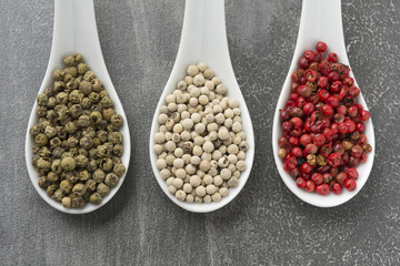 Three white ceramic spoons filled with various peppers on rustic grey background
