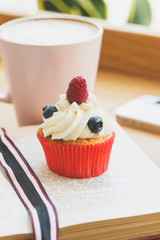 Small cupcake with raspberry and blueberry on the book, cup of hot cappuccino