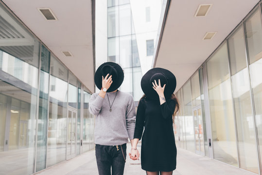 Portrait of young couple in urban environment, holding hands, covering faces with hats