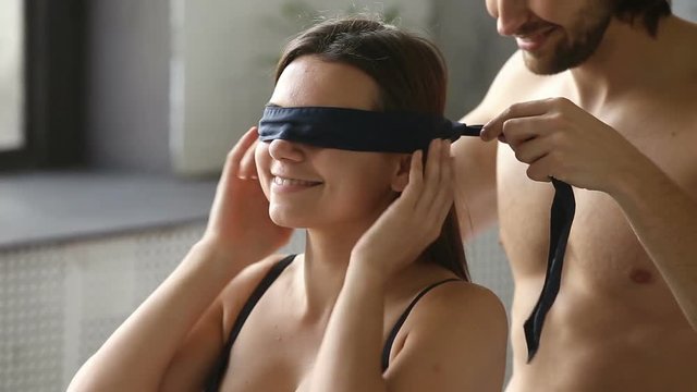 Young man blindfolding smiling woman sitting on bed, couple enjoy foreplay playing erotic role game with bdsm fetish black ribbon on eyes, husband embracing kissing wife before making love having sex