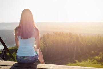 Rear view of a seated girl in a blue T-shirt and looking at the mountains and the forest in the distance