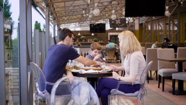 A young family dines at a restaurant, a woman is photographed by her husband, who drinks tea, their child drinks a milkshake from a straw