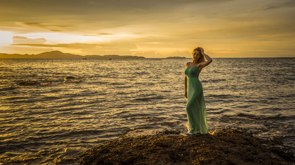 A beautiful sexy blonde in a long turquoise dress stands on the seashore against a backdrop of a...