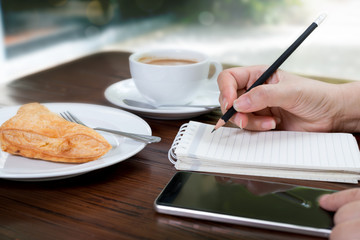 Business woman start a new day with fresh idea coffee and pie.Woman writing note with pencil and using smart phone during simple breakfast at home.