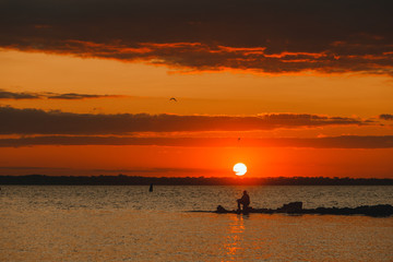 Fototapeta premium Fisherman standing on a pier at dawn sky background with sun rays and reflected in the sea water. Silhouette of fishermen boats fishing in sunset at the beach.