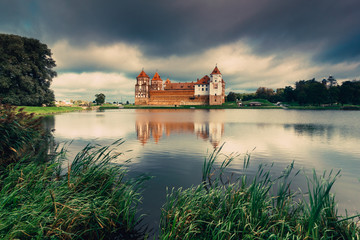 Obraz premium View of Mir Castle - ancient medieval fortress on the shore of the lake, reflecting in the water. Dramatic cloudy sky. Architectural monument of the Belarus. UNESCO World Heritage. Vintage effect. 