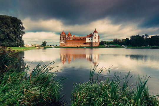 View of Mir Castle - ancient medieval fortress on the shore of the lake, reflecting in the water. Dramatic cloudy sky. Architectural monument of the Belarus. UNESCO World Heritage. Vintage effect. 