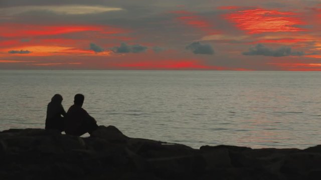 Sunset by the ocean. Calm water.Couple sitting on the beach and admiring the sunset. Silhouette