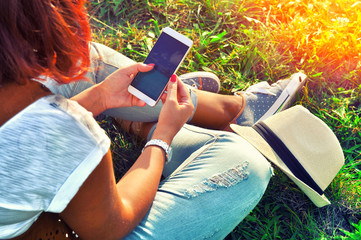 Relaxation with a mobile. Break time. Young woman using cellphone and sitting on the grass wearing...