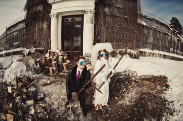 A beautiful ginger bride white fur coat groom in colored cheerful glasses shape of heart are playing on dope and rake in the snowy park on building and wood background. Winter wedding outdoors.