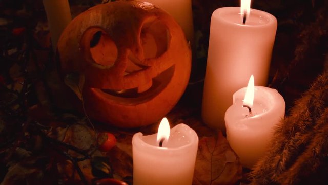 All Saints' Day, Thanksgiving, Halloween, Harvest Festival. Little one pumpkin with a carved cheerful face lies on the autumn maple leaves and smiles.Large candles,small candles are burning around.